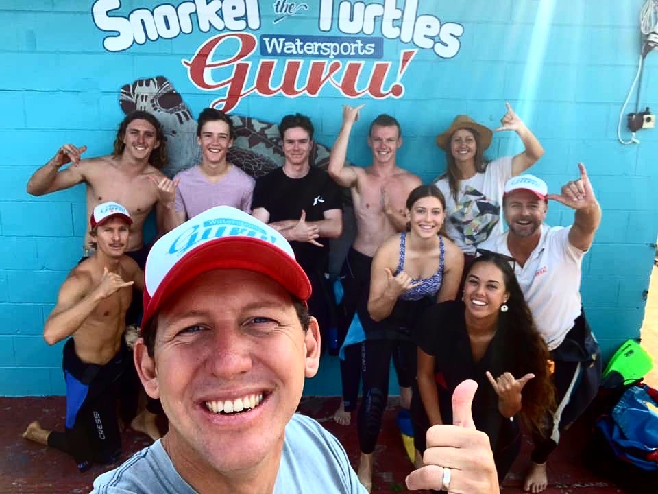 Group of happy snorkelers posing in front of a "Snorkel with Turtles" Watersports Guru sign